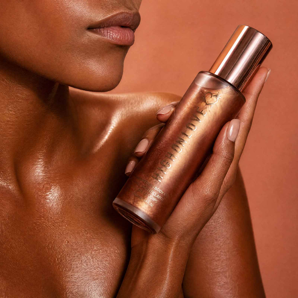 High On Love Shimmering Body Oil held by woman with glowing bronze shimmer skin against warm terracotta background showing luminous body glow finish